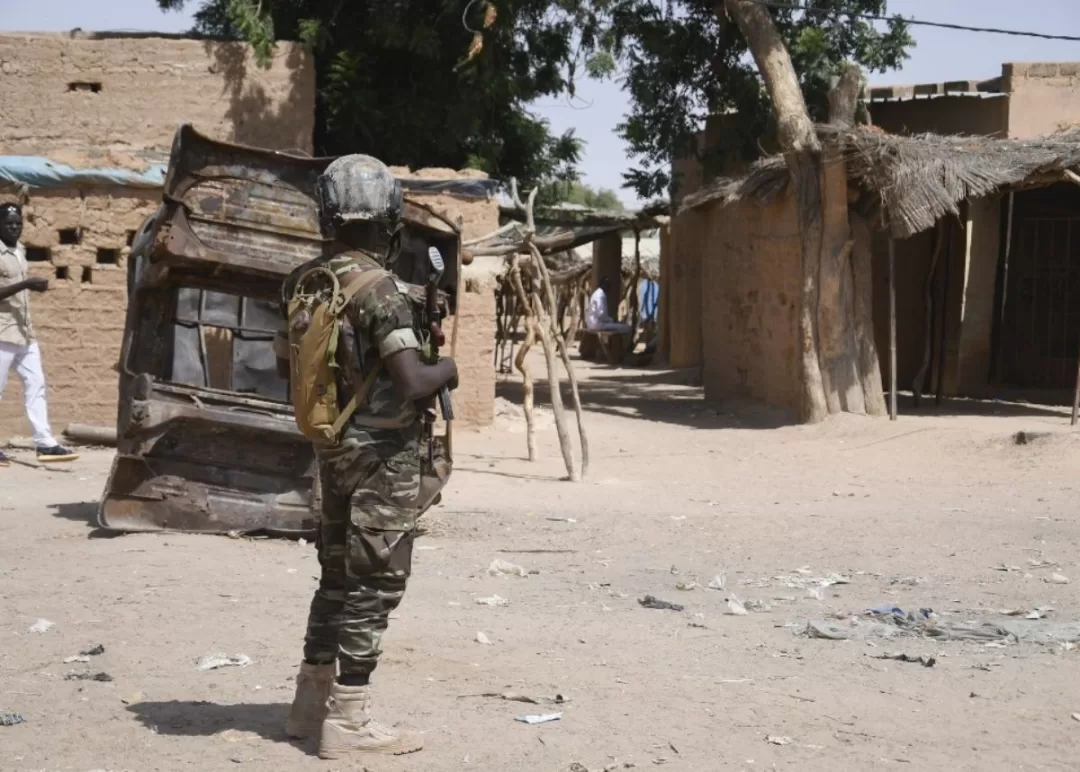 Militaire en tenue camouflée et casque tenant une arme dans un village désert avec bâtiments en terre et arbres autour