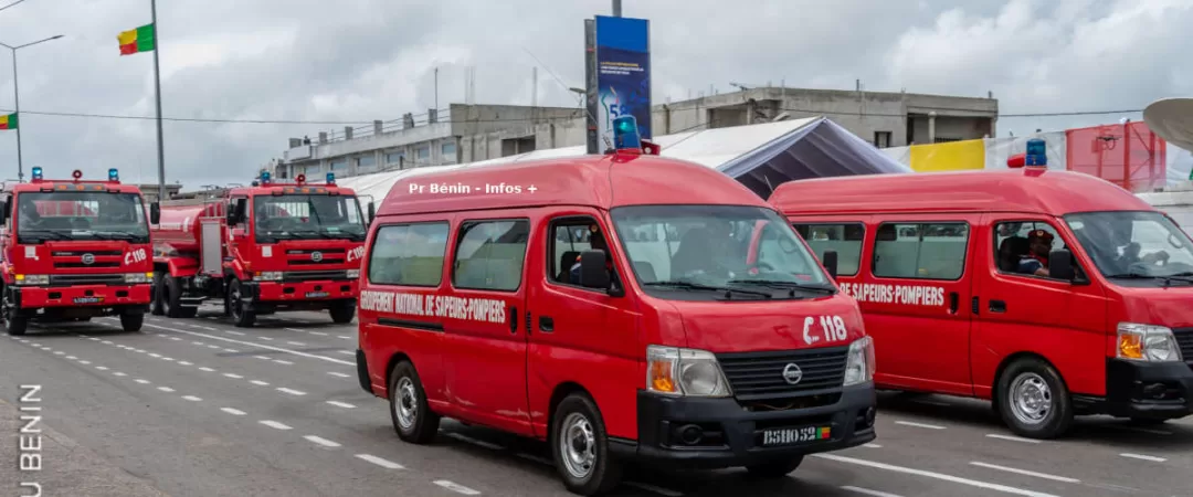 Plusieurs véhicules rouges de sapeurs-pompiers alignés sur une route avec un drapeau en arrière-plan sous un ciel nuageux