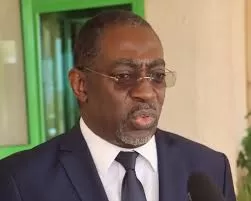 Homme portant un costume sombre, chemise blanche et cravate noire parlant devant un micro dans un cadre intérieur simple