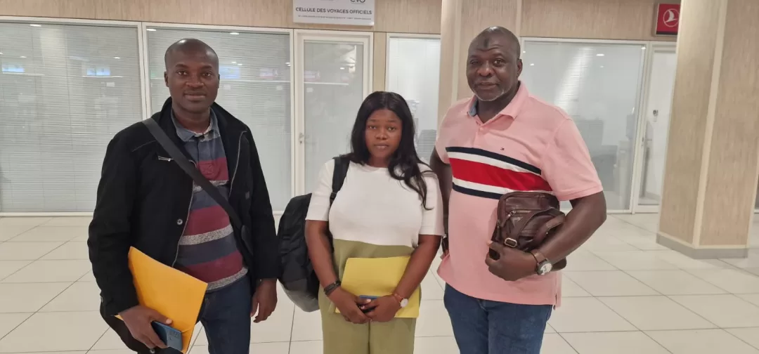 Trois personnes souriantes posent dans un hall intérieur avec des dossiers jaunes à la main et un sac à dos noir visible