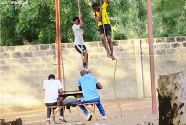 Deux adolescents grimpent à la corde tandis que deux adultes assis à une table semblent noter ou observer l'épreuve sportive