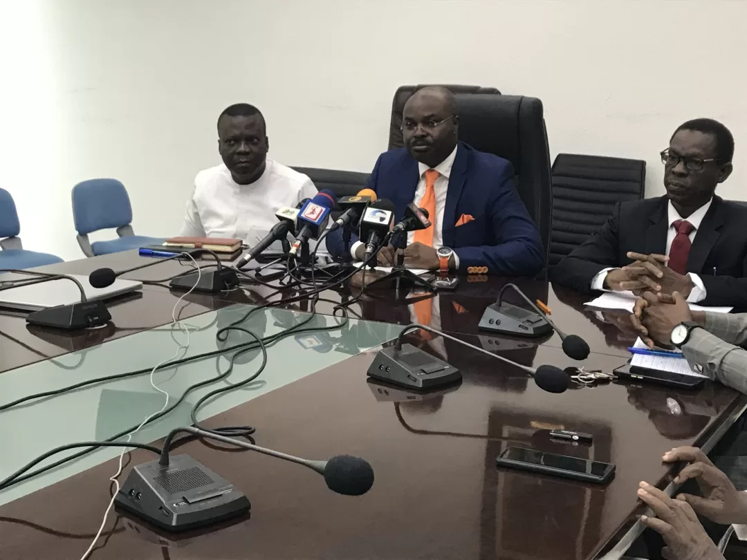 Trois hommes assis à une table de conférence avec plusieurs microphones et enregistreurs devant eux
