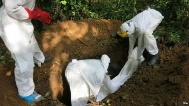 Trois personnes en combinaison de protection enterrent un corps dans une fosse creusée en pleine nature