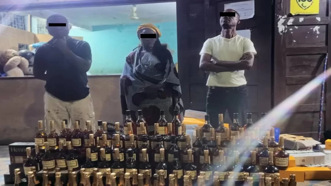 Trois individus masqués debout derrière une grande quantité de bouteilles d'alcool empilées sur une table devant une porte ouverte