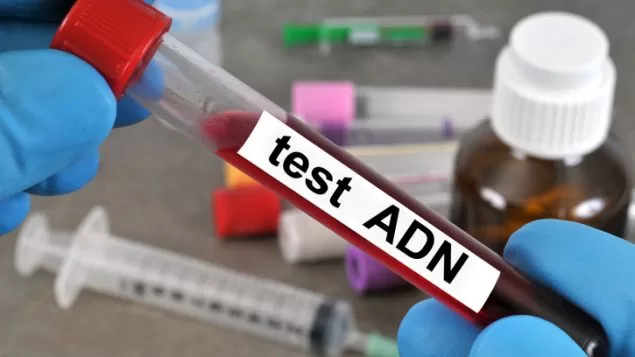 Une main gantée tient un tube rempli de sang étiqueté test ADN avec seringues et flacons en arrière-plan