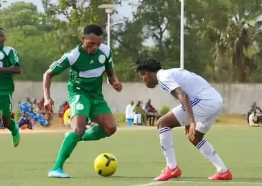 Deux joueurs de football en action sur un terrain gazonné devant des spectateurs assis au bord du terrain
