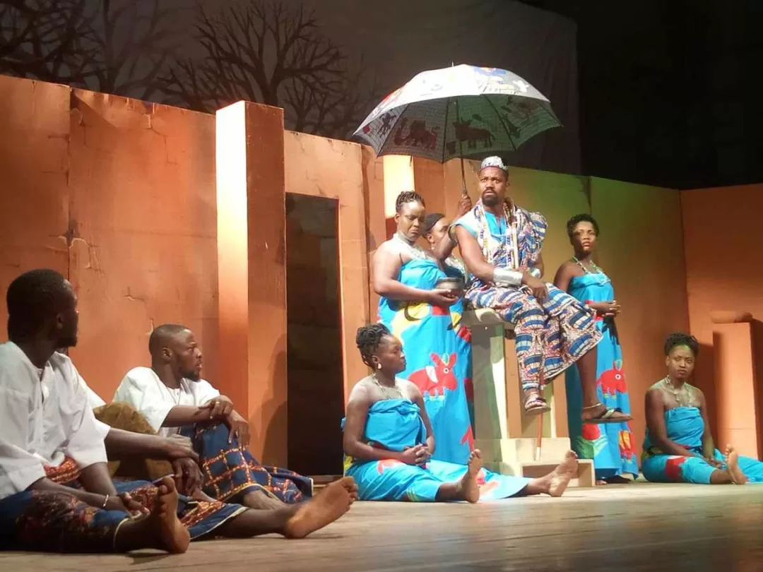 Des artistes en costume traditionnel sont assis et debout sur une scène avec un décor évoquant un village africain.