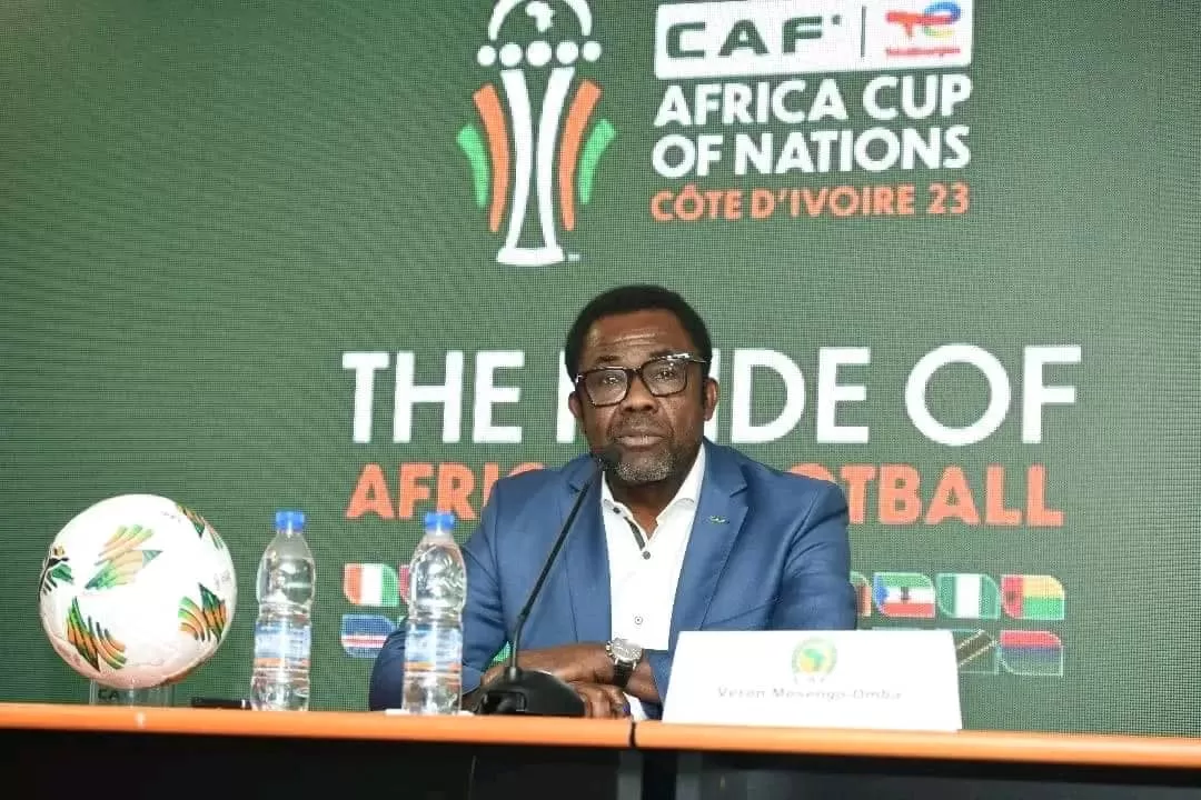 Homme assis derrière une table avec micro, devant un écran affichant le logo de la CAN Côte d’Ivoire 2023 et une balle de football posé à côté de bouteilles d’e