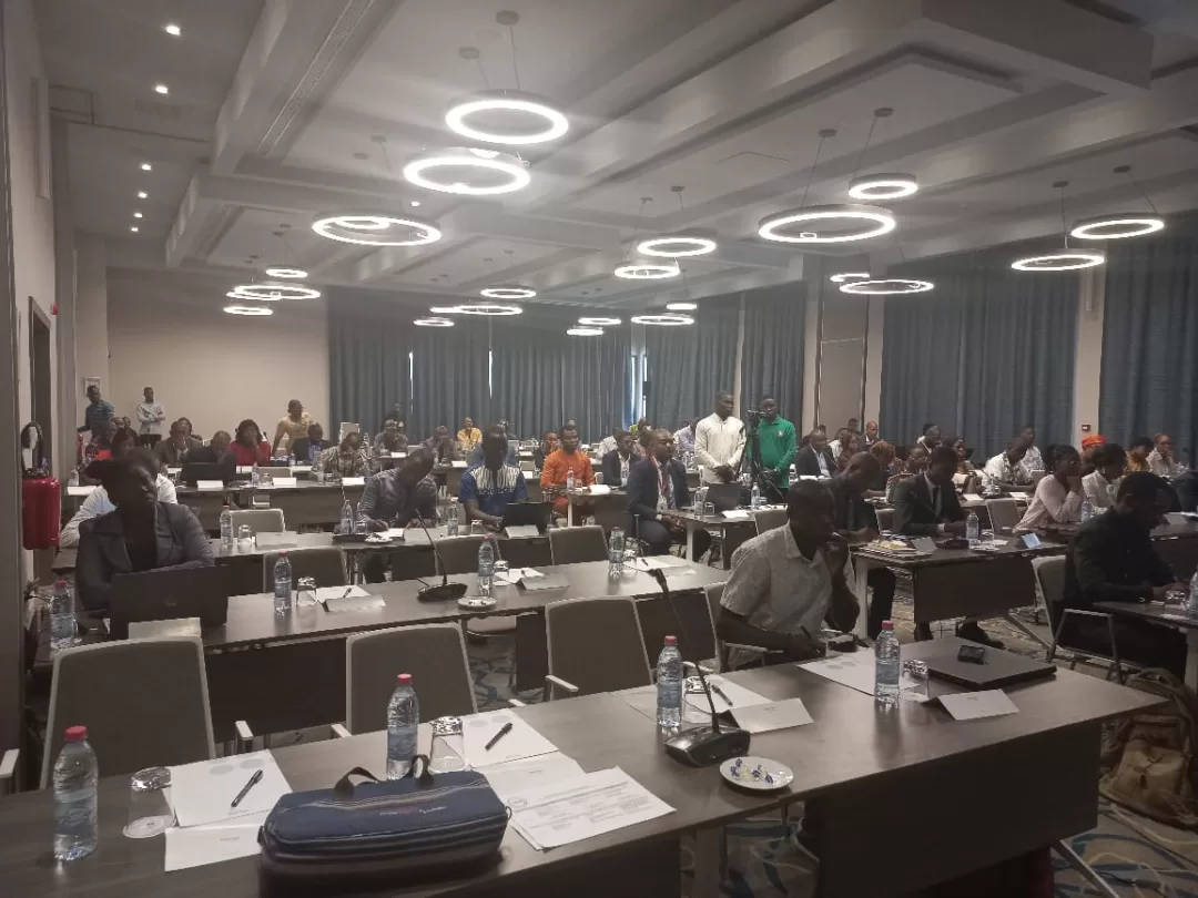 Salle de conférence avec des participants assis à des tables équipées de documents, bouteilles d'eau et micros sous des luminaires ronds modernes