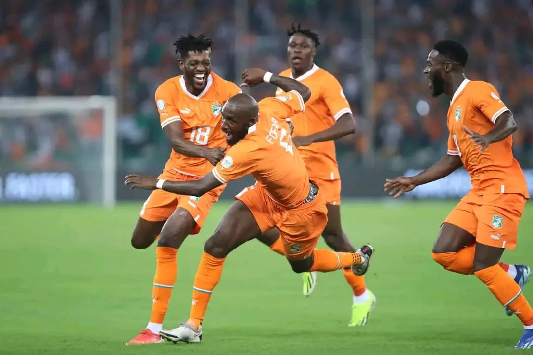 Quatre joueurs en maillot orange célébrant un but sur un terrain de football devant un public nombreux