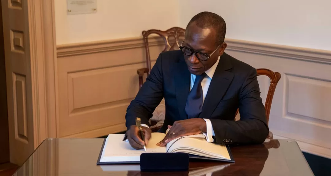 Homme en costume sombre assis à une table en bois signe un grand livre ouvert dans une pièce aux murs clairs
