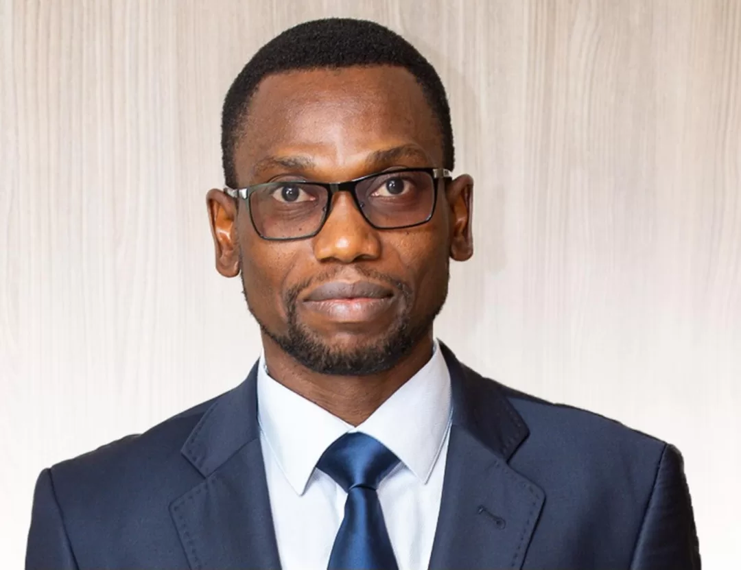 Homme portant un costume sombre, une chemise blanche et une cravate bleue, avec des lunettes noires devant un mur clair