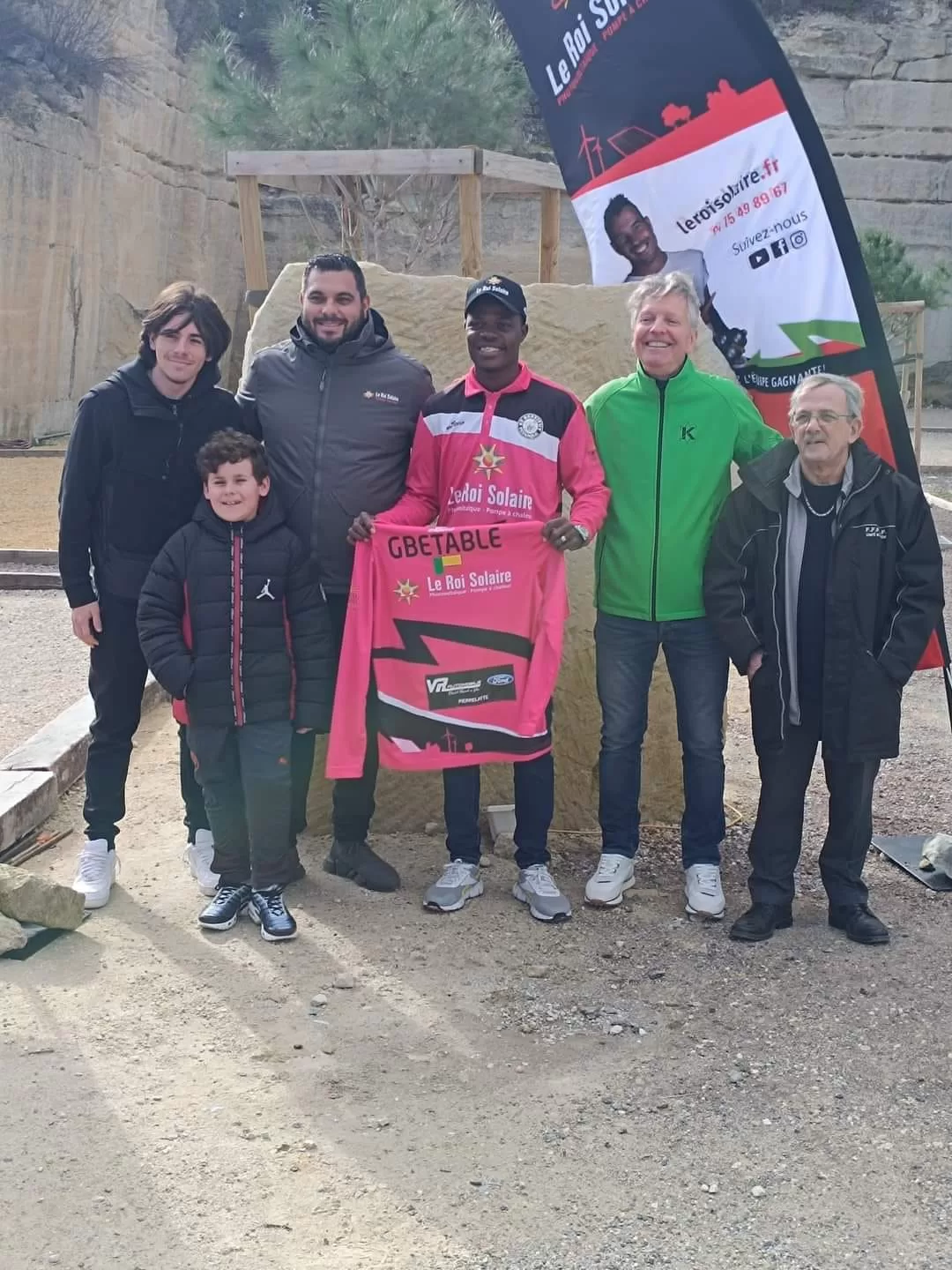 Six hommes posent debout sur un terrain avec un maillot rose tenu par l’homme central devant un panneau publicitaire.