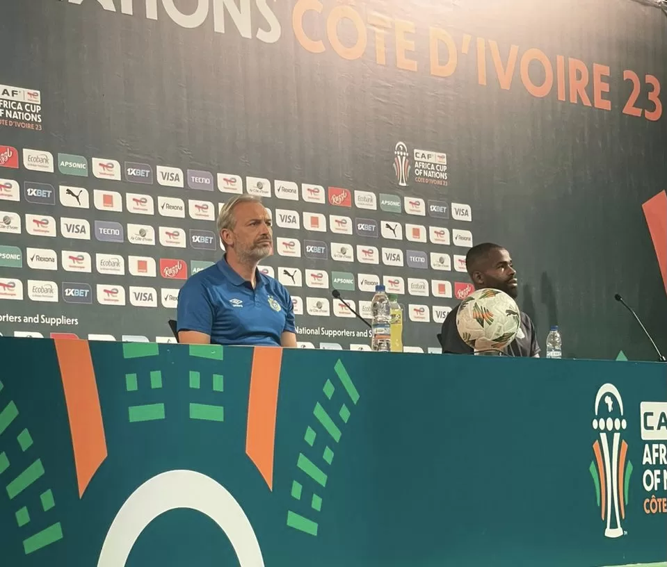 Deux hommes assis à une table de conférence avec un ballon de football et un fond décoré pour une compétition sportive africaine.