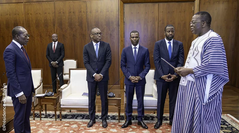 Cinq hommes en costume et un homme en tenue traditionnelle debout dans une salle avec des fauteuils et un tapis rouge