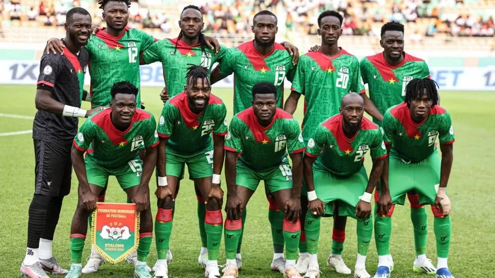 Onze joueurs en maillot vert avec détails rouges posant ensemble sur un terrain de football avant un match
