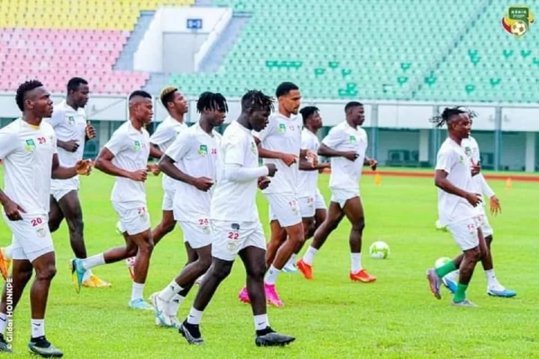 Des joueurs de football en tenue d’entraînement blanc courent sur une pelouse dans un stade aux gradins multicolores
