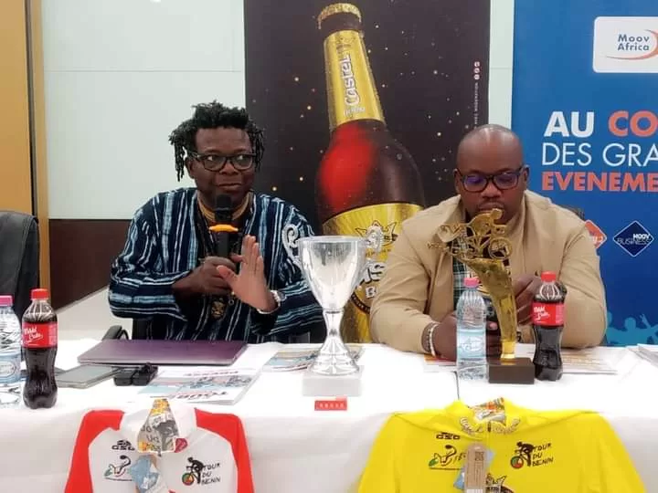 Deux hommes assis à une table avec des trophées, des maillots de cyclisme et des bouteilles de boisson devant eux