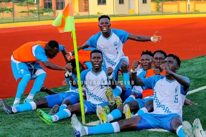 Six joueurs de football en maillots bleu et blanc célèbrent assis et allongés près d'un drapeau de coin sur un terrain en herbe synthétique verte