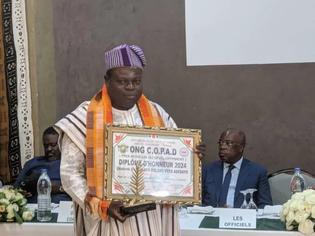 Un homme en tenue traditionnelle tient un diplôme encadré et un trophée avec des personnes assises derrière lui à une table.