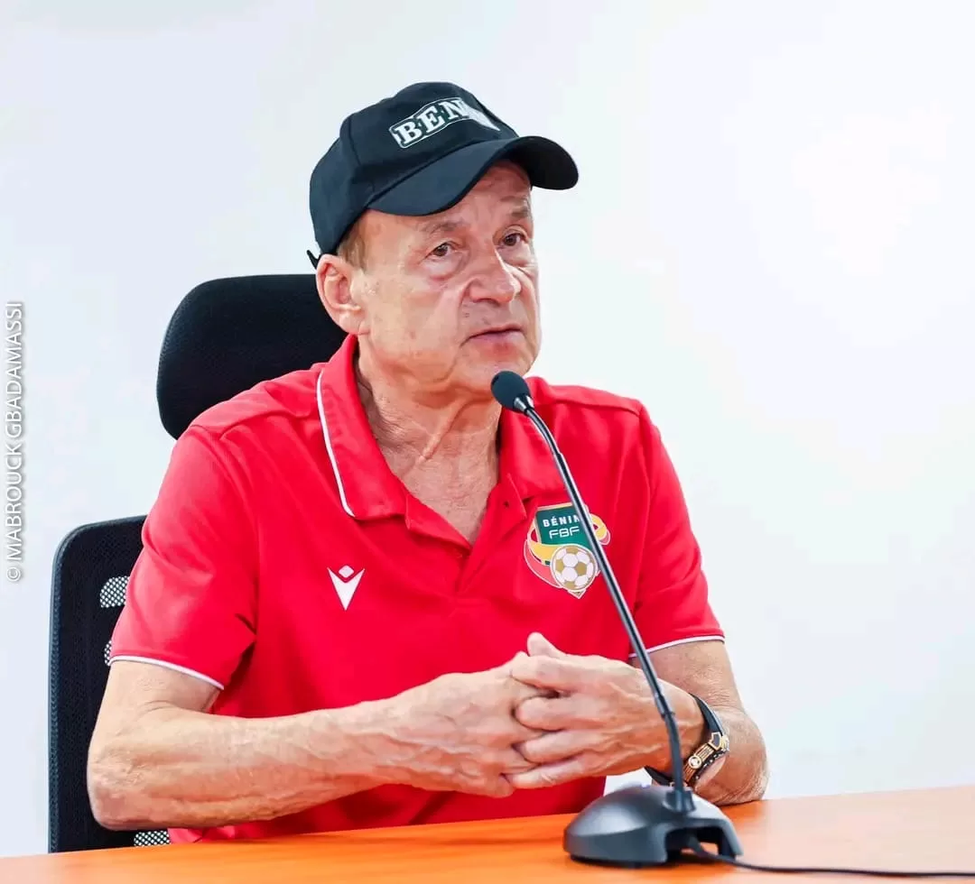 Homme portant une casquette et un polo rouge avec un blason, parlant devant un micro posé sur une table en bois claire