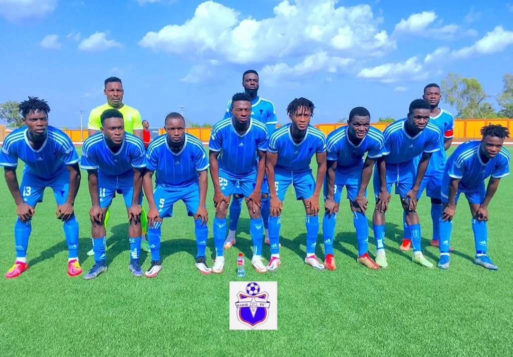 Onze joueurs de football en tenue bleue posent sur un terrain vert sous un ciel bleu avec quelques nuages blancs