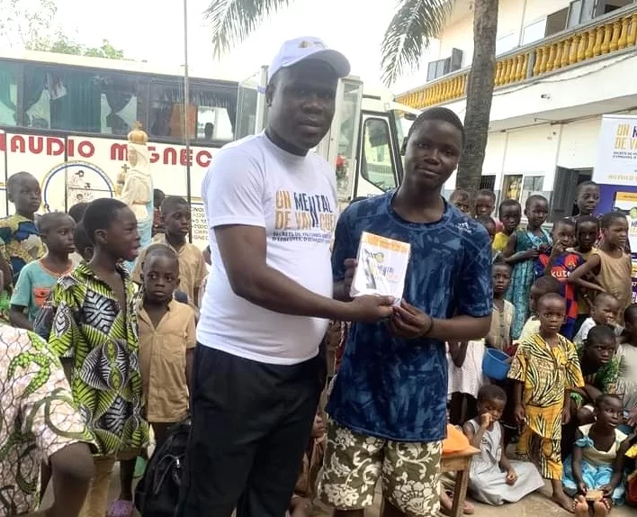 Un homme en casquette blanche remet un livre à un adolescent devant un groupe d’enfants rassemblés à l’extérieur