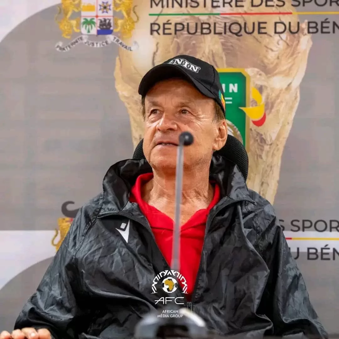 Homme assis en veste noire et casquette, micro devant lui, devant un panneau officiel du ministère des sports