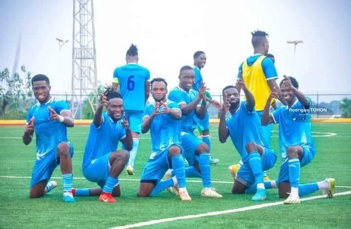 Joueurs en tenue bleue célébrant à genoux et debout sur un terrain de football gazonné sous un ciel clair