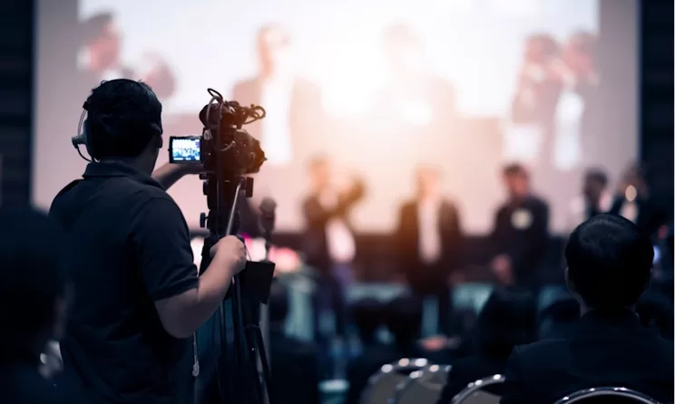 Une personne filme une conférence où plusieurs intervenants sont assis devant un public dans une salle obscure