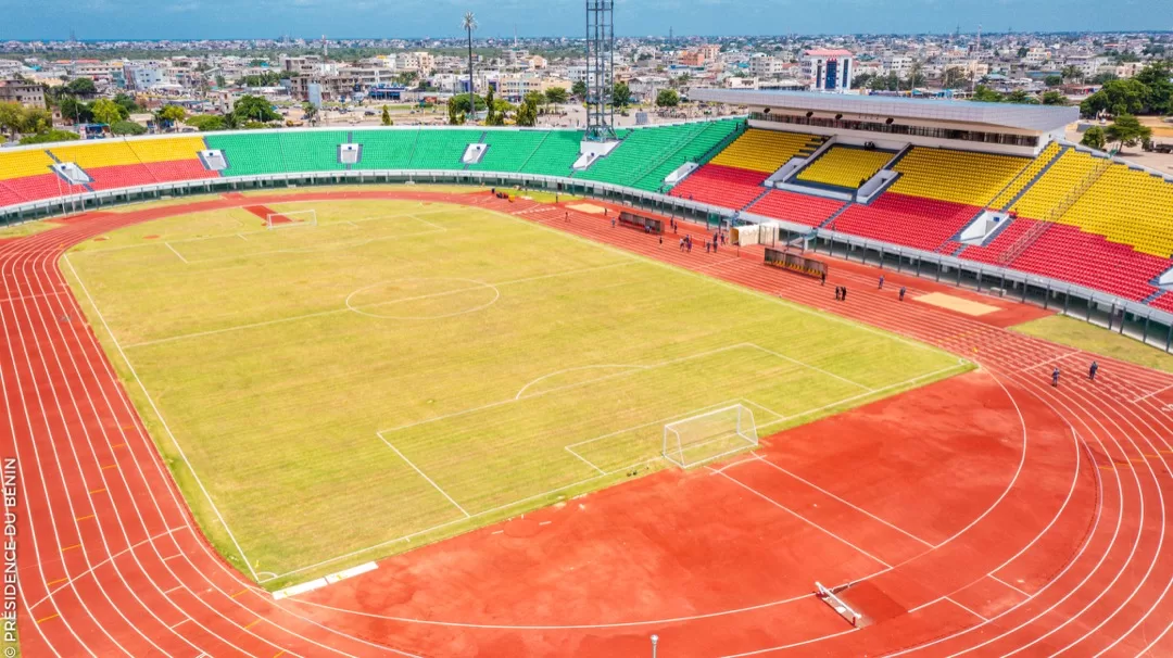 Stade avec pelouse partiellement verte et gradins aux couleurs vives entourés d’une piste d’athlétisme rouge et vue urbaine en arrière-plan