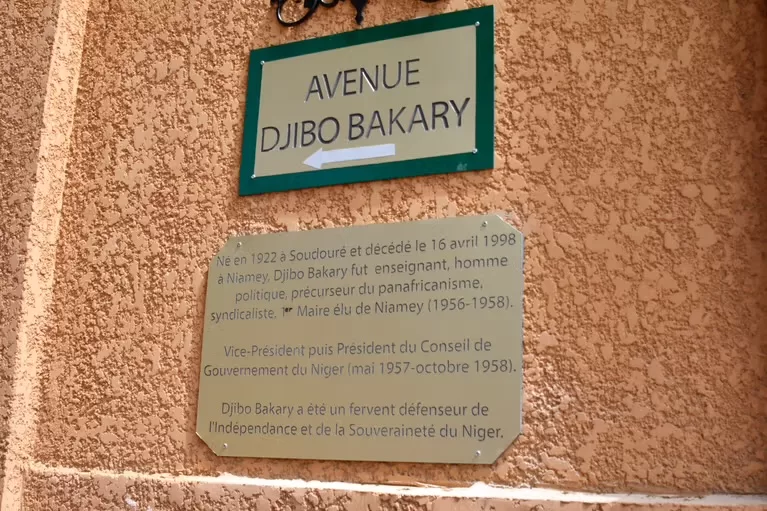 Plaque de rue verte indiquant Avenue Djibo Bakary avec une plaque explicative sur un mur ocre