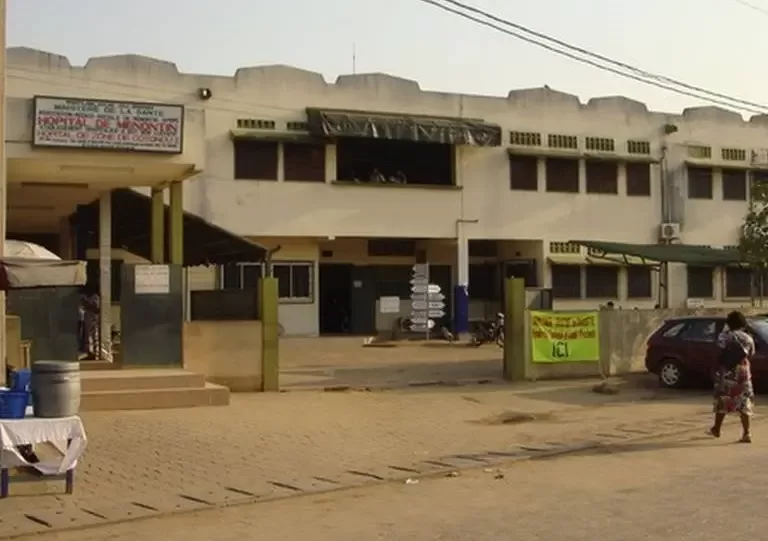 Façade claire d’un bâtiment à deux étages avec entrée, panneaux et véhicules stationnés devant une personne debout
