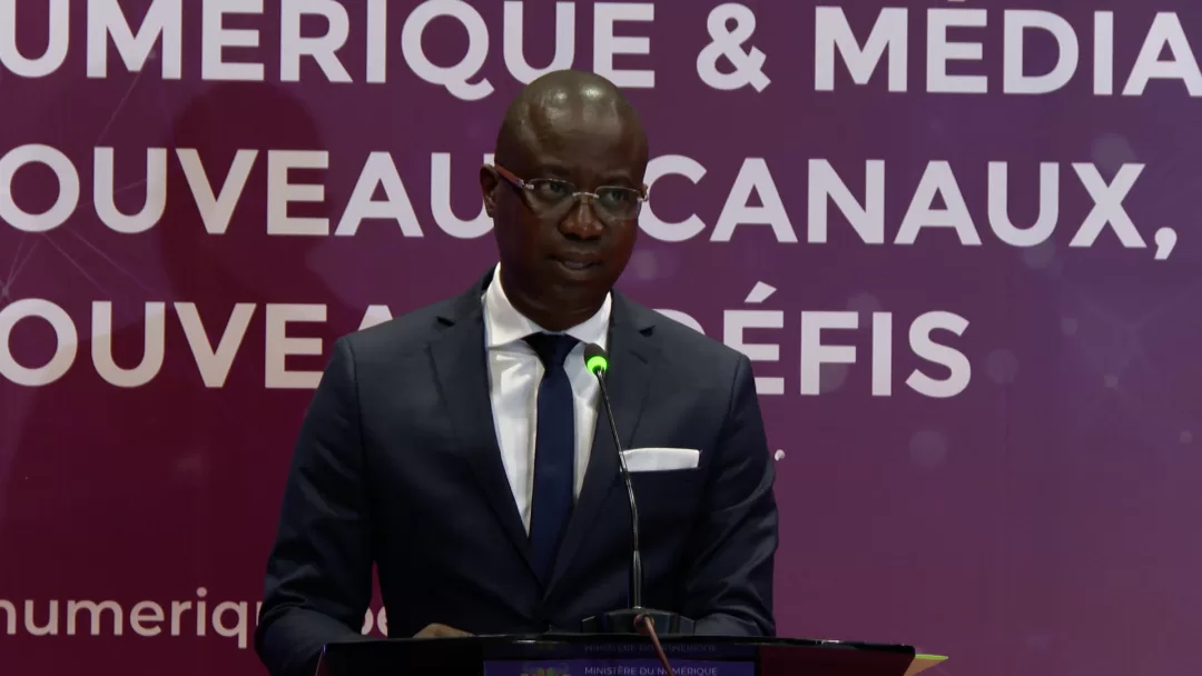 Un homme en costume sombre parle derrière un pupitre devant un fond textuel sur le numérique et les médias