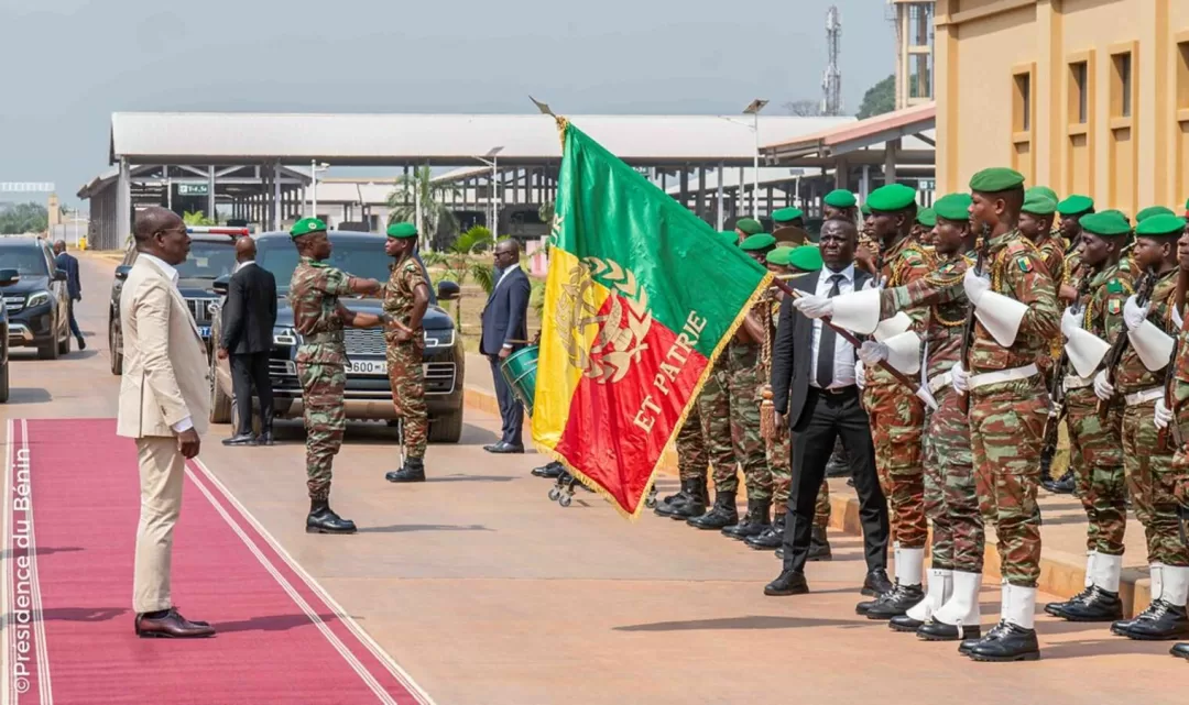 Un homme en costume clair salue une formation de soldats en tenue camouflage et bérets verts avec un drapeau tricolore sur un tapis rouge extérieur