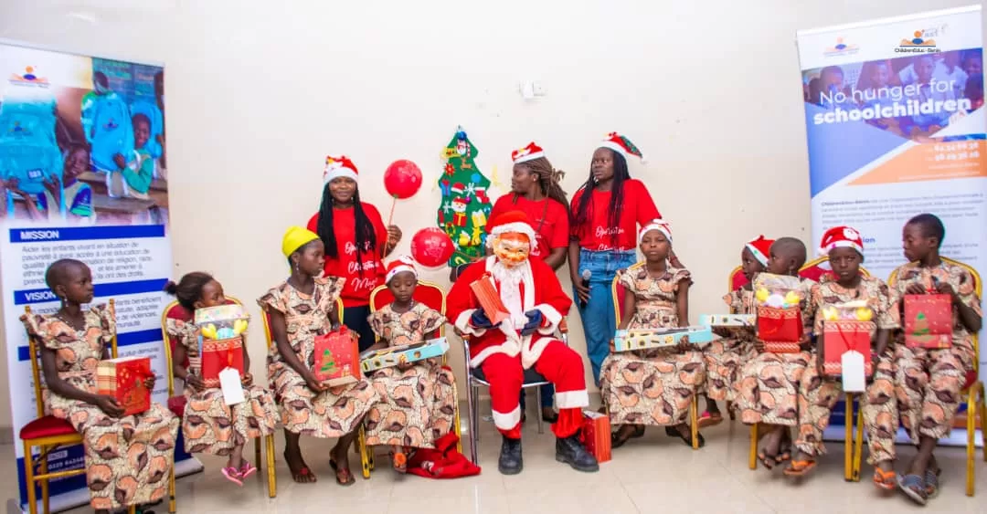 Un groupe d’enfants en tenue assortie et plusieurs adultes avec chapeaux de Noël posent autour d’un Père Noël et d’un sapin décoré