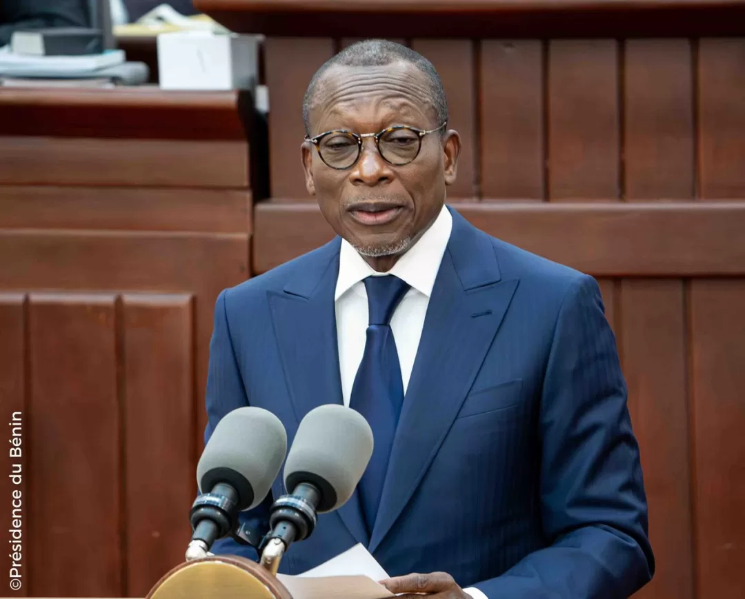 Homme en costume bleu et lunettes parlant devant deux micros dans un cadre officiel en bois brun