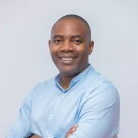 Un homme souriant en chemise bleue claire, bras croisés, posant devant un fond uni gris clair