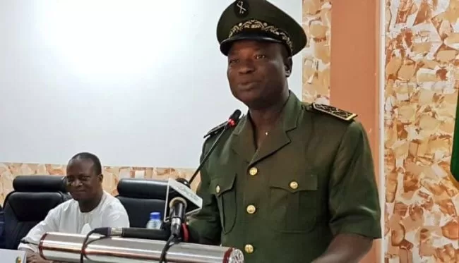 Un homme en uniforme militaire parle derrière un pupitre avec un microphone, un autre homme est assis en arrière-plan