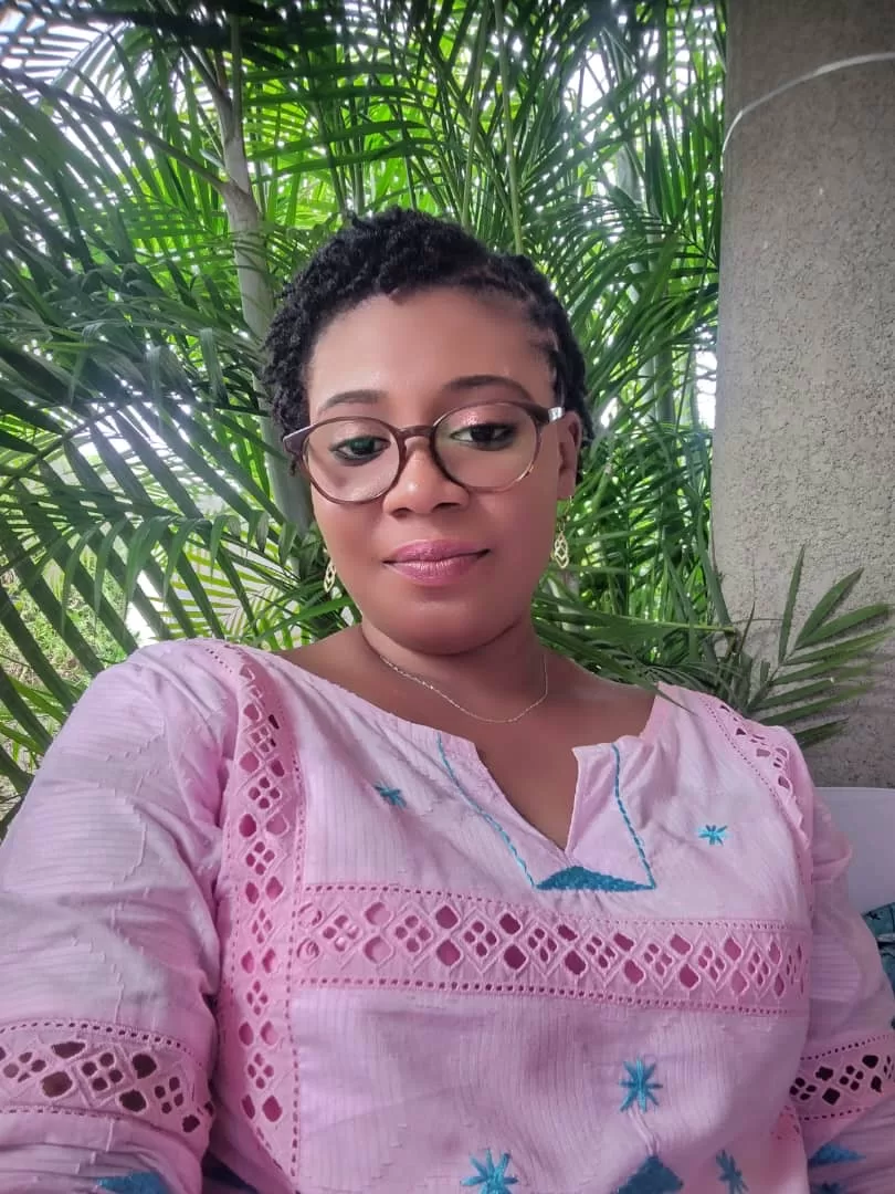 Femme portant des lunettes et un vêtement brodé rose avec un fond de végétation verte et un mur gris