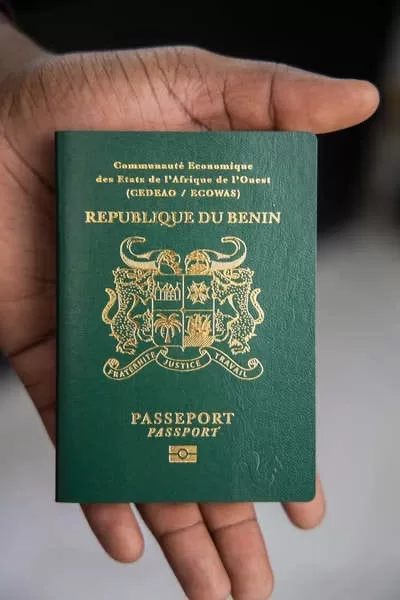 Une main tient un passeport vert foncé avec inscriptions dorées "République du Bénin" et un blason au centre