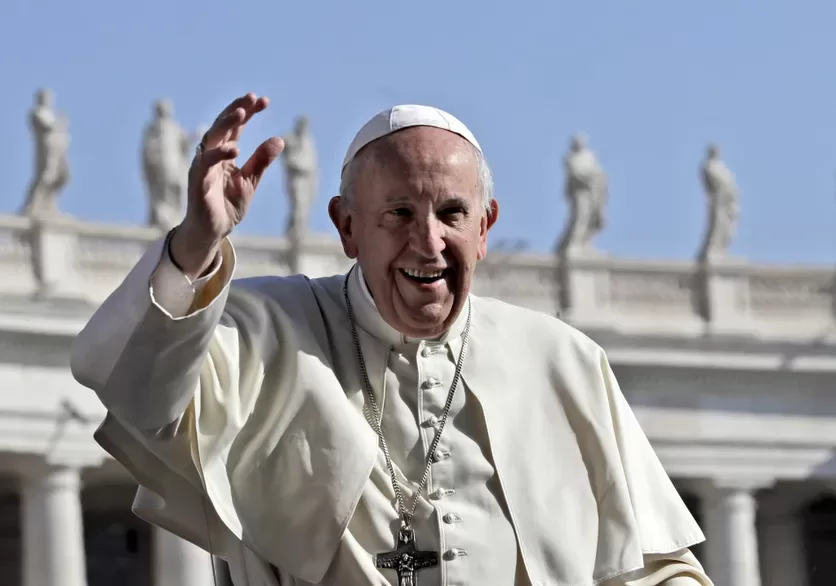 Un homme en tenue religieuse blanche agite la main devant un bâtiment décoré de statues sous un ciel bleu