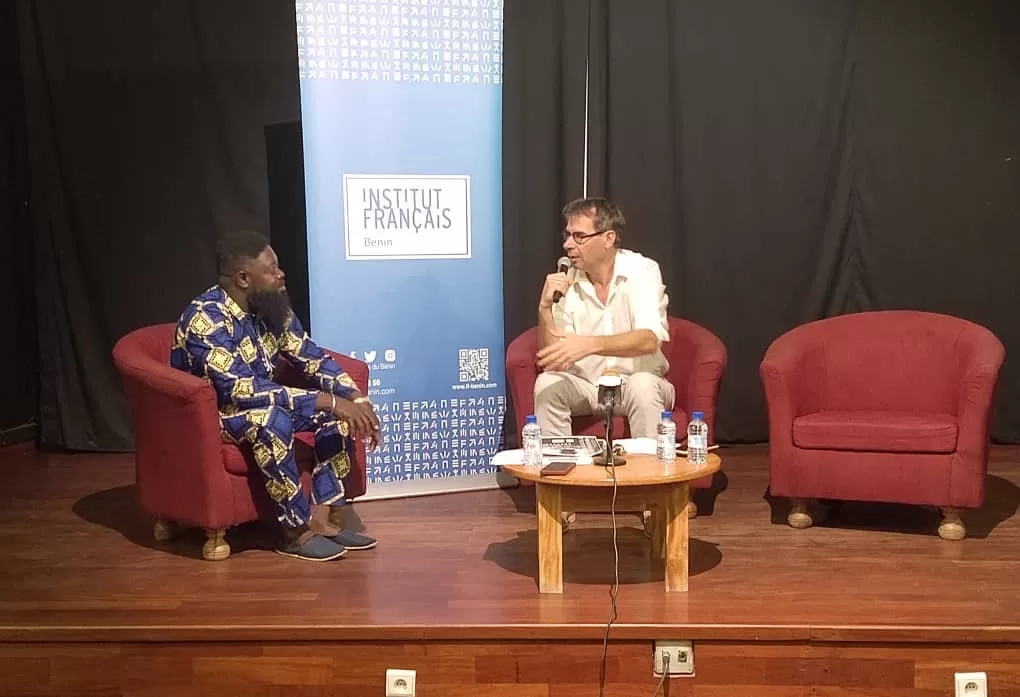 Deux hommes assis sur des fauteuils rouges sur une scène discutent face à un roll-up bleu de l'Institut Français et une petite table en bois devant eux