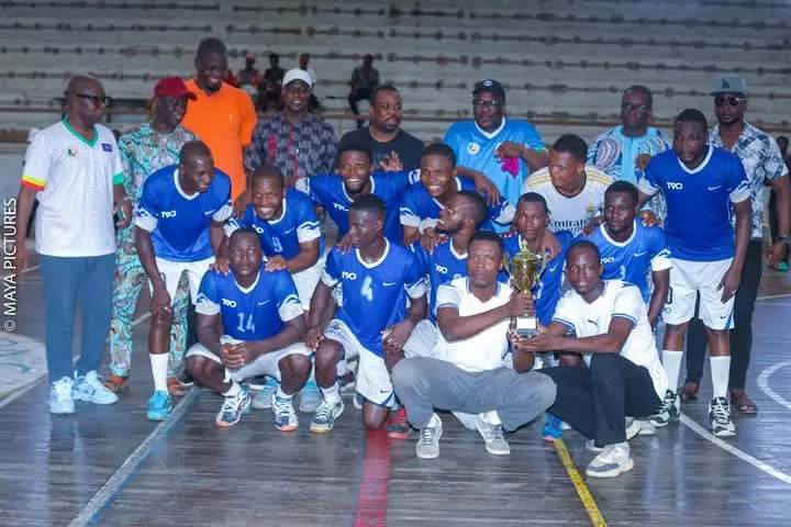 Équipe de joueurs en tenue bleue posant avec un trophée au milieu d’un terrain couvert avec des personnes debout derrière eux