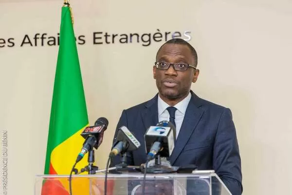 Un homme en costume sombre et lunettes parle devant plusieurs micros avec un drapeau vert, jaune et rouge en arrière-plan