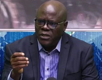 Homme portant des lunettes noires et costume sombre parlant en gesticulant avec la main droite devant un micro