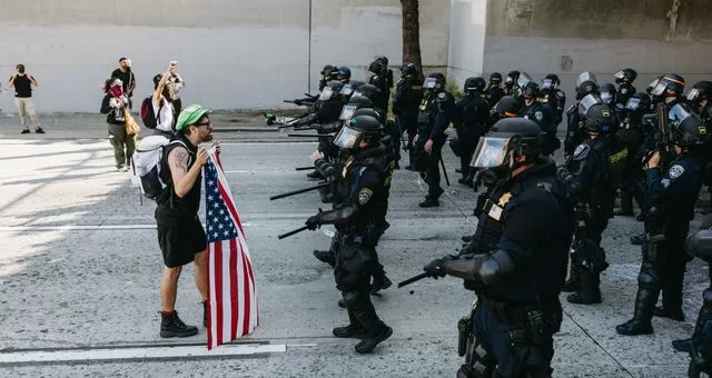Un manifestant tenant un drapeau américain fait face à une ligne de policiers en tenue anti-émeute sur une rue vide