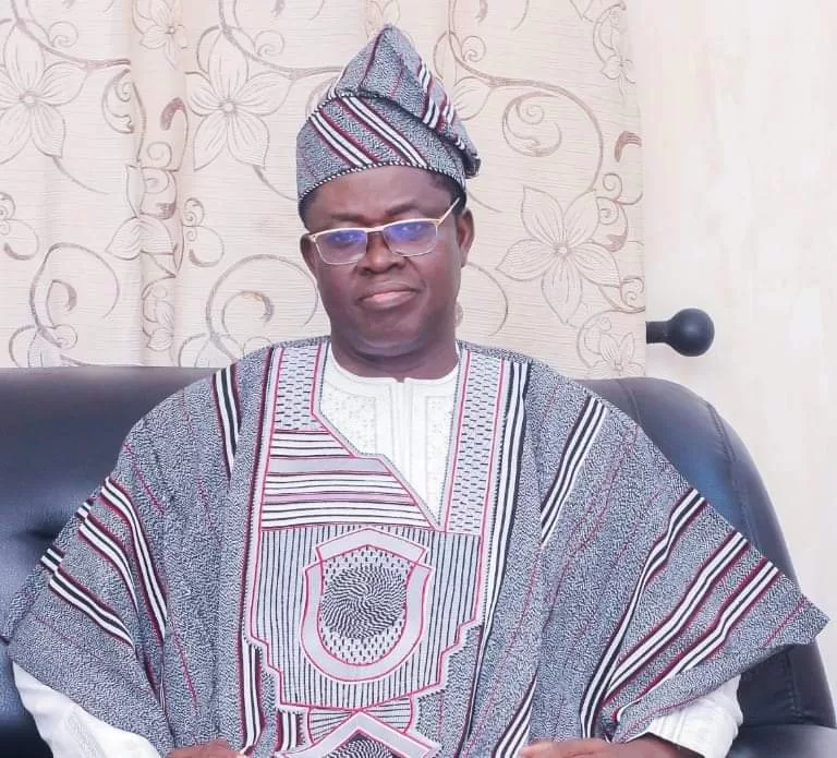 Homme assis sur un canapé en tenue traditionnelle africaine rayée avec chapeau assorti et lunettes devant un rideau fleuri beige