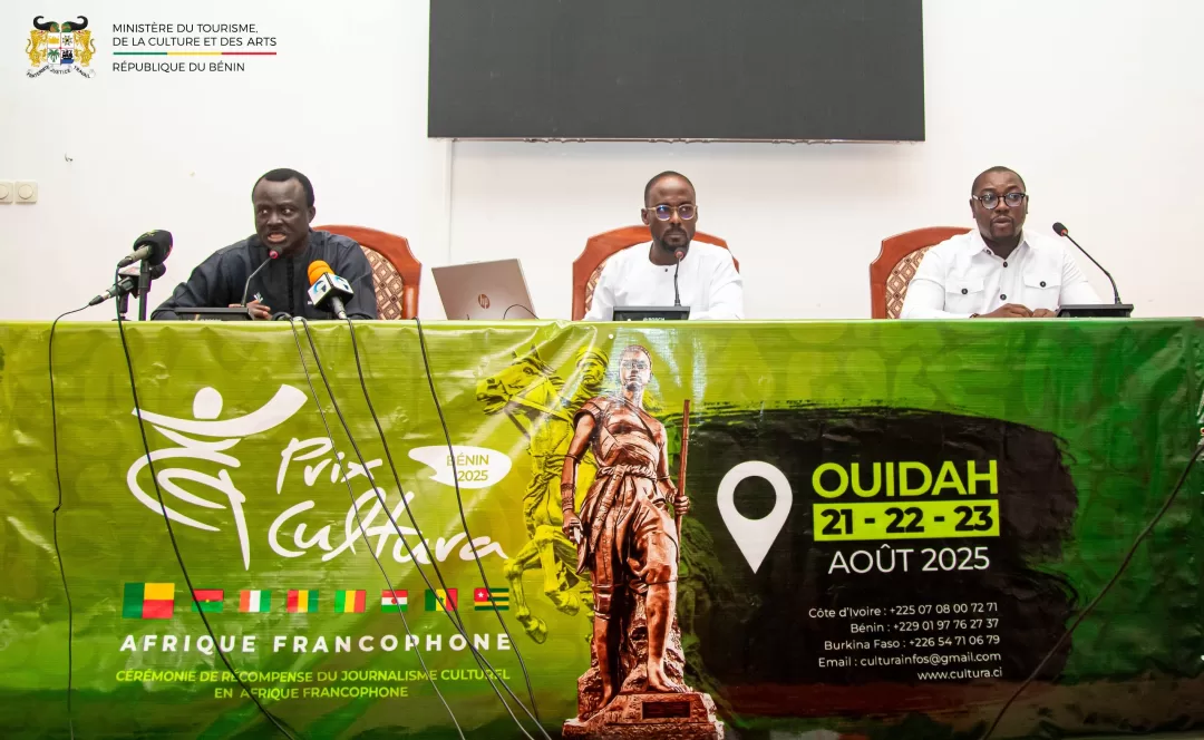 Trois hommes assis derrière une table verte avec micro, devant une bannière annonçant un événement en août 2025 à Ouidah