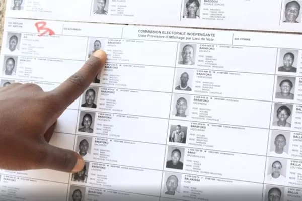 Main masculine pointant une ligne sur une liste électorale affichée avec photos et informations des électeurs